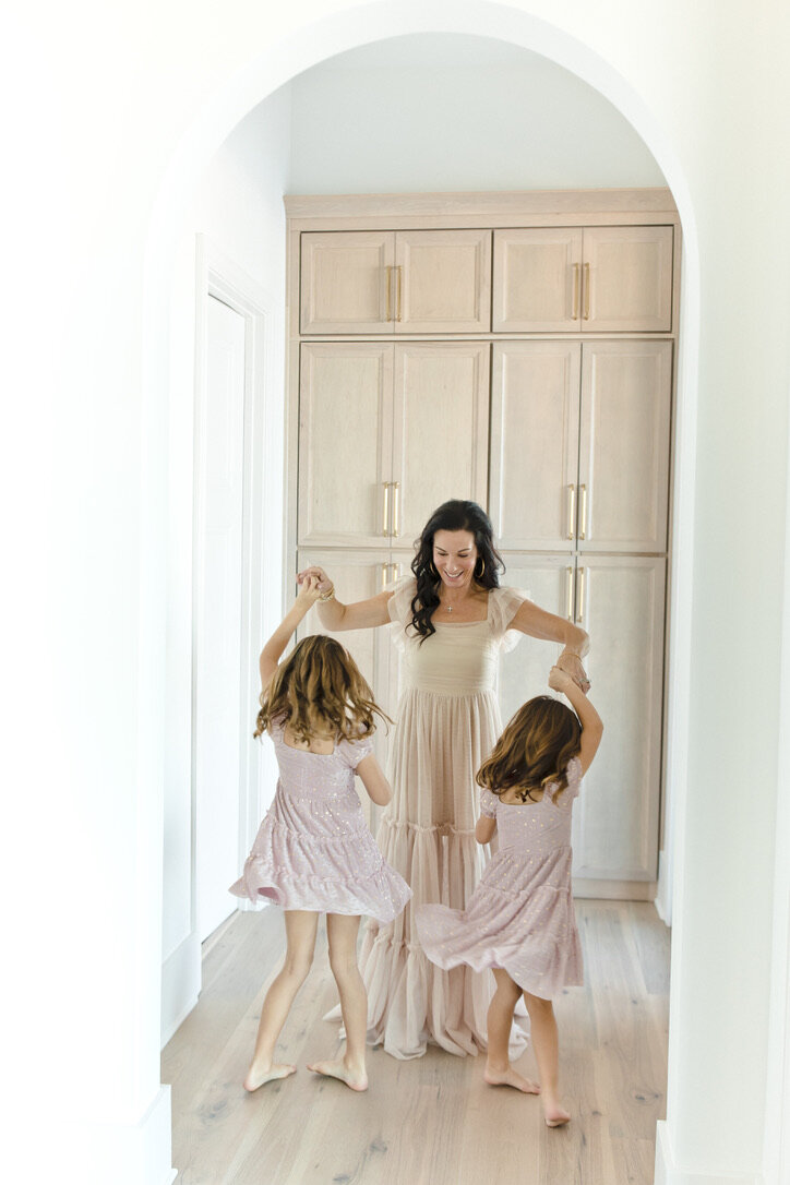 Mom dancing with her two young daughters in a sunlit hallway, a joyful moment that holds the grief in motherhood of watching them grow